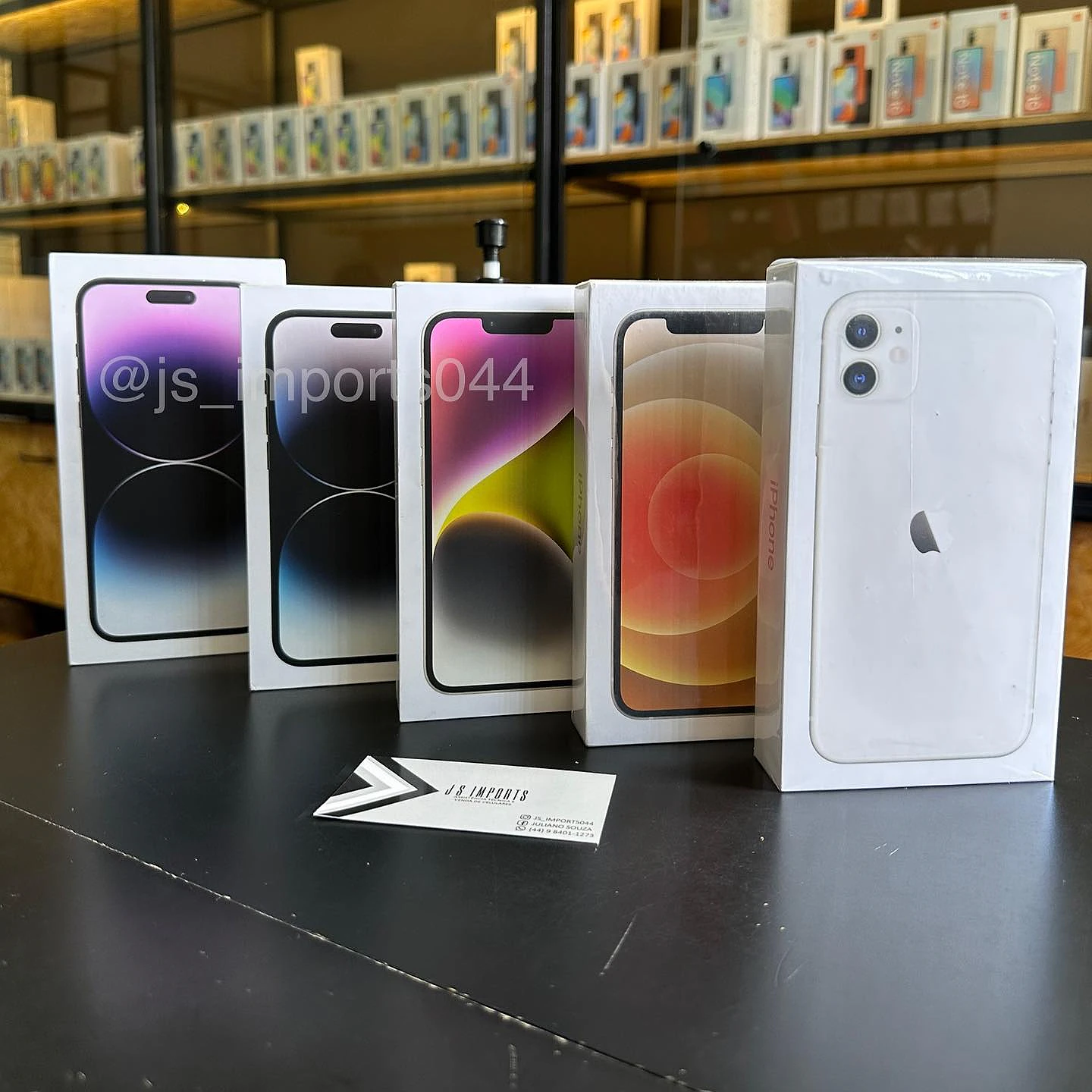 Caixas de iPhone lacradas em pé no balcão da JS Imports, com o cartão da loja na frente